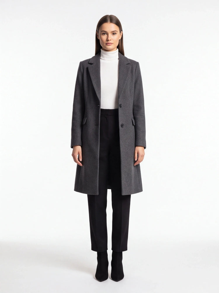 A woman stands against a plain white studio background wearing a long dark gray overcoat over a white turtleneck, black pants, and black ankle boots.