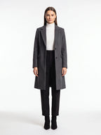 A woman stands against a plain white studio background wearing a long dark gray overcoat over a white turtleneck, black pants, and black ankle boots.