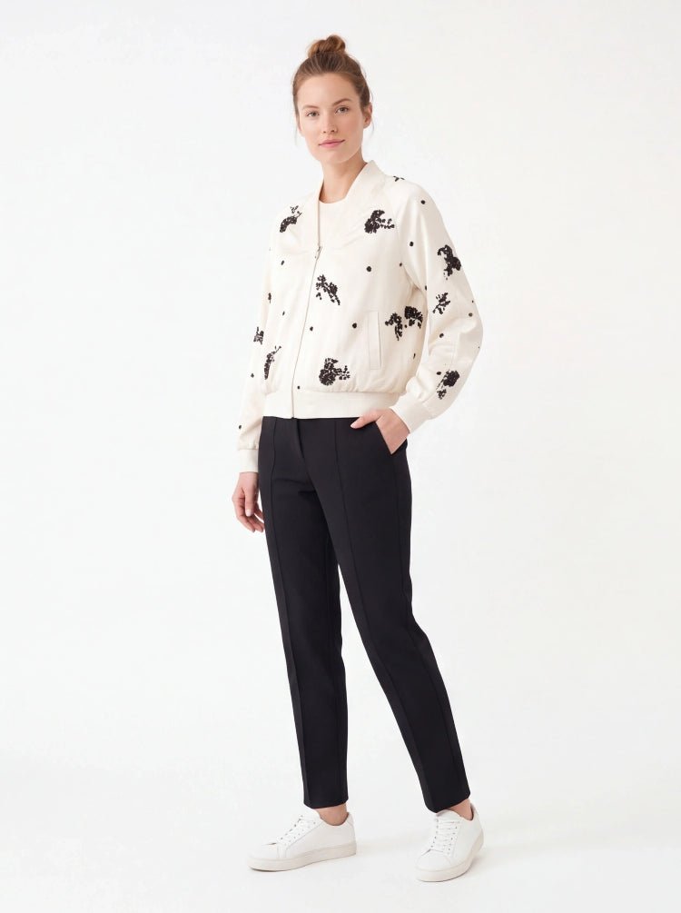A woman modeling a cream-colored bomber jacket with black embroidered motifs, paired with black trousers and white sneakers, set against a plain white studio background.