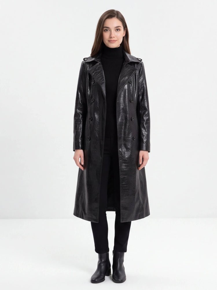 A woman modeling a long black faux-leather trench coat over a black turtleneck, black pants, and black ankle boots, photographed against a seamless white studio background.