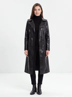 A woman modeling a long black faux-leather trench coat over a black turtleneck, black pants, and black ankle boots, photographed against a seamless white studio background.