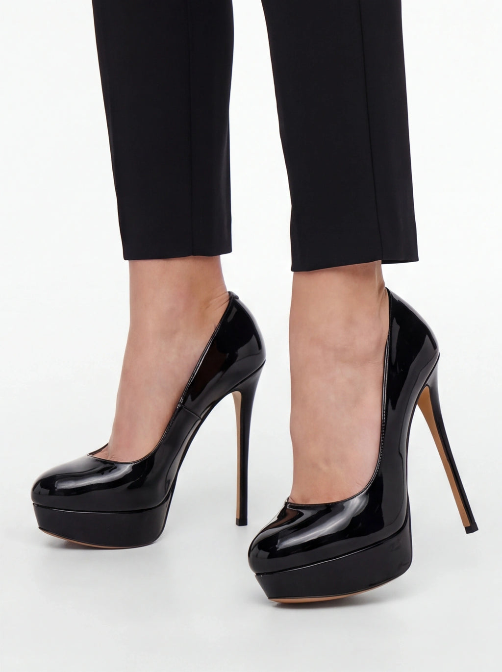 Black patent leather platform high heels with stiletto heels, shown on a model wearing black cropped trousers against a light gray studio background.