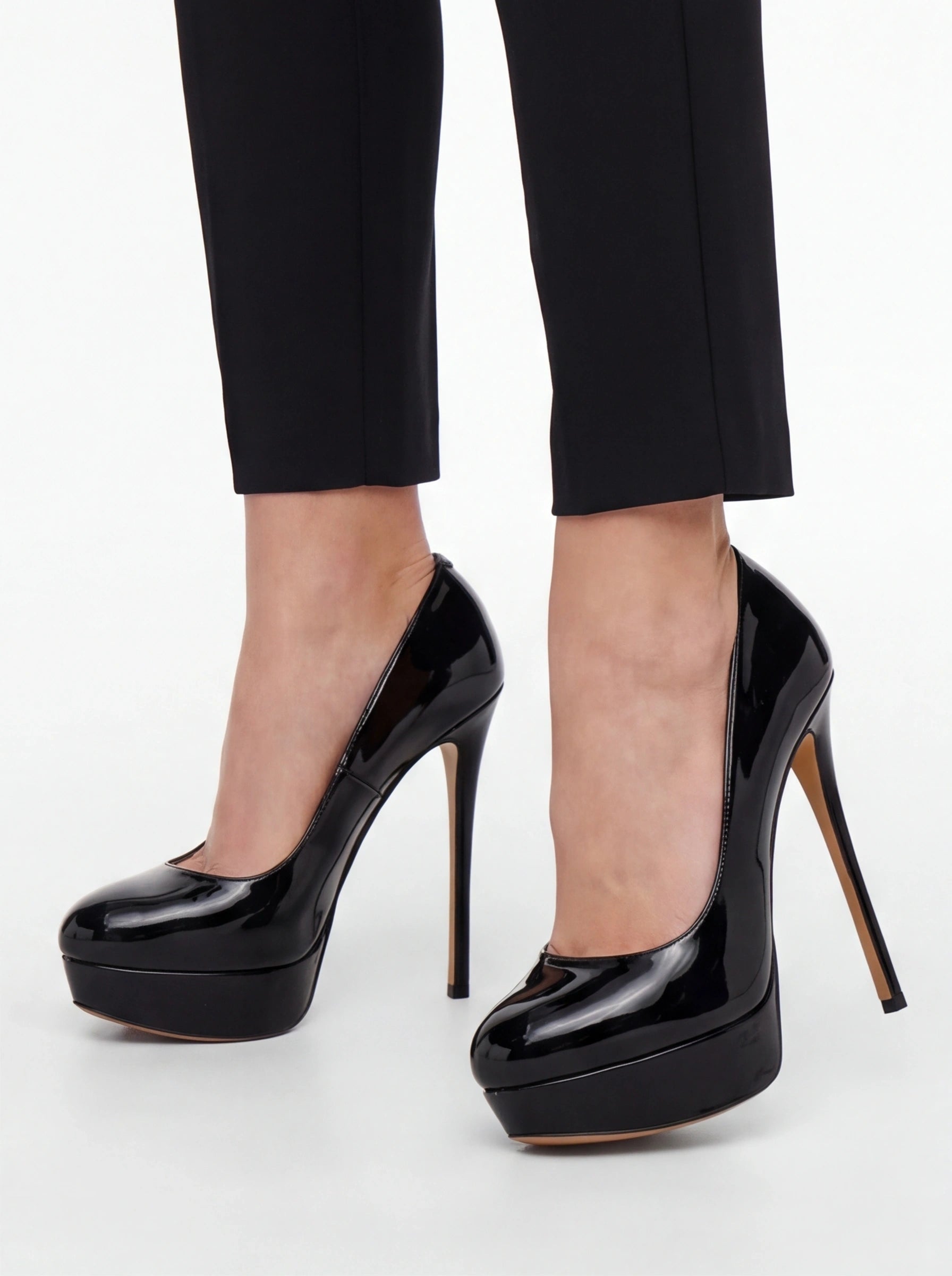 Black patent leather platform high heels with stiletto heels, shown on a model wearing black cropped trousers against a light gray studio background.