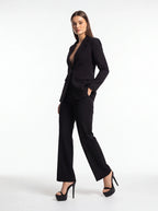 A woman modeling a black tailored pantsuit with a structured blazer and wide-leg trousers, paired with black high-heeled sandals, against a plain white studio background.