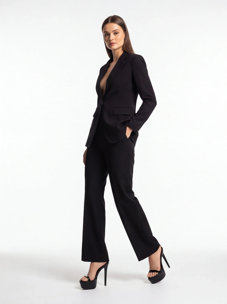 A woman modeling a black tailored pantsuit with a structured blazer and wide-leg trousers, paired with black high-heeled sandals, against a plain white studio background.