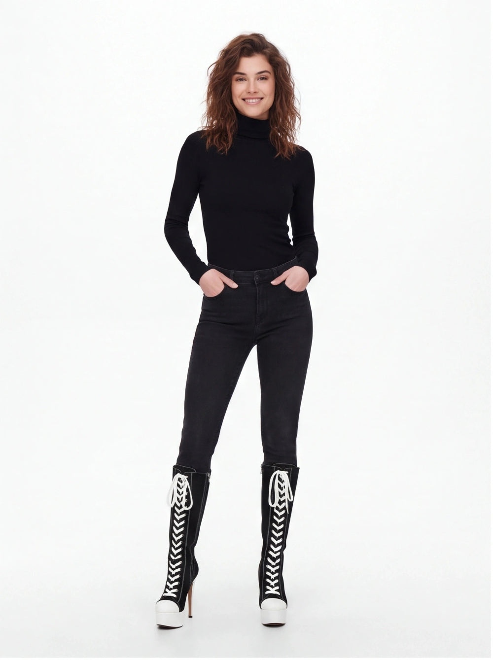 A woman modeling a black long-sleeve turtleneck top, black skinny jeans, and black lace-up knee-high boots with white laces and white platform heels, against a plain white studio background.