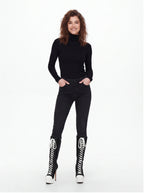 A woman modeling a black long-sleeve turtleneck top, black skinny jeans, and black lace-up knee-high boots with white laces and white platform heels, against a plain white studio background.