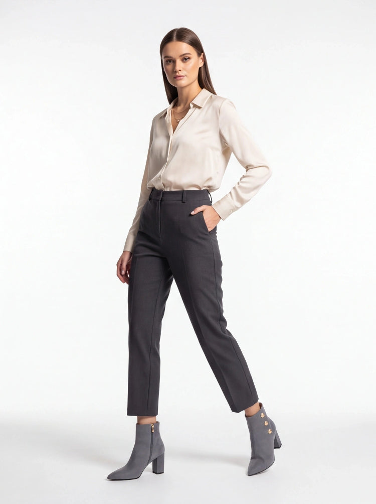 A woman modeling a light beige satin button-down shirt, dark charcoal gray cropped trousers, and gray heeled ankle boots against a plain white studio background.