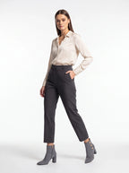 A woman modeling a light beige satin button-down shirt, dark charcoal gray cropped trousers, and gray heeled ankle boots against a plain white studio background.