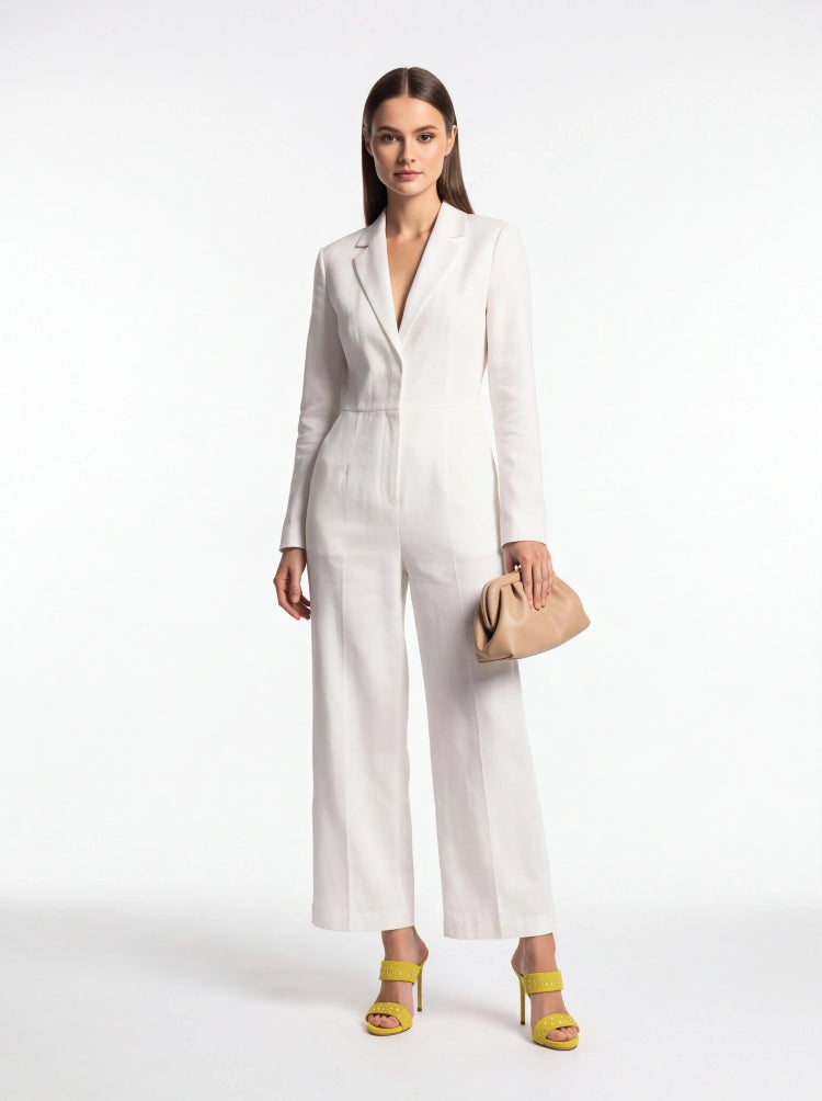 A woman modeling a white long-sleeve wide-leg jumpsuit with a deep V-neckline, paired with yellow heeled sandals and a beige clutch bag, set against a plain white studio background.