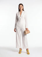 A woman modeling a white long-sleeve wide-leg jumpsuit with a deep V-neckline, paired with yellow heeled sandals and a beige clutch bag, set against a plain white studio background.