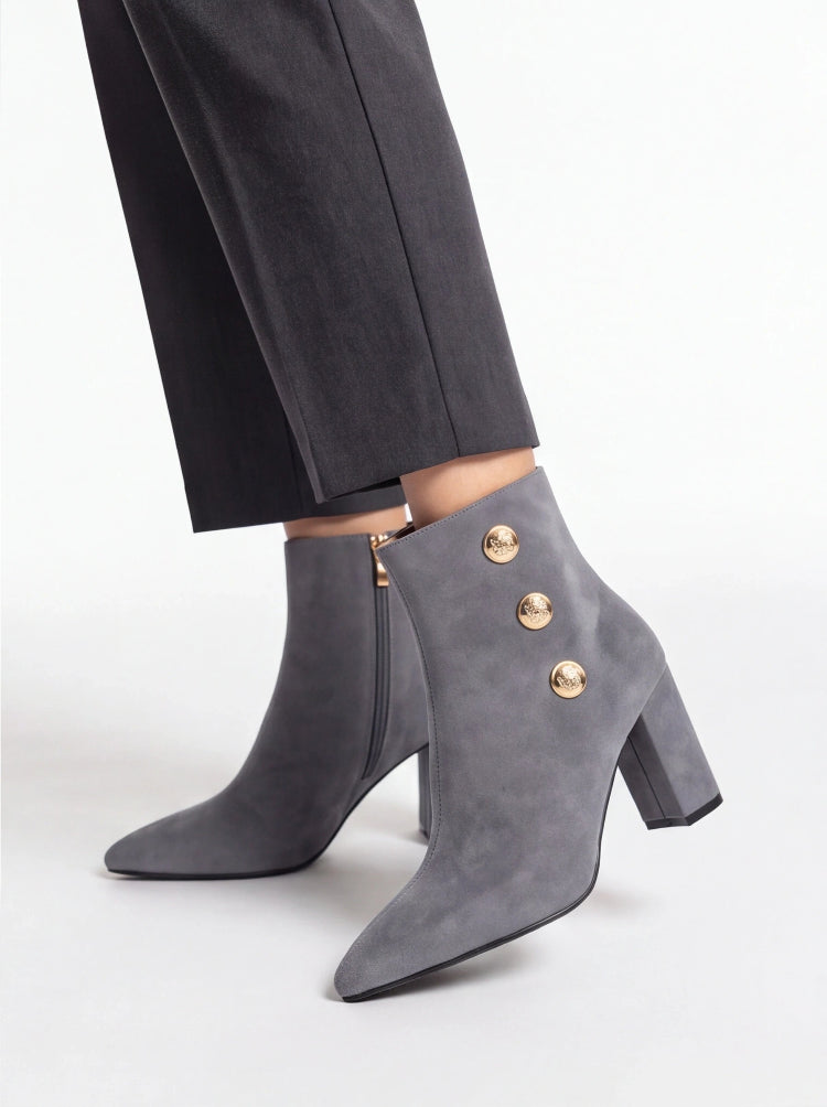 Gray suede ankle boots with a block heel, pointed toe, and three gold-tone decorative buttons on the outer side, shown worn with dark gray trousers against a light gray studio background.