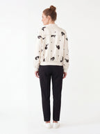 A woman shown from the back wearing a cream-colored bomber jacket with black embroidered cow patterns, paired with black trousers and white sneakers, against a plain white studio background.