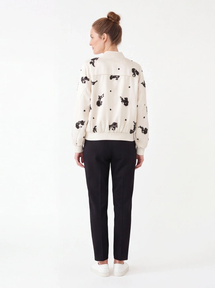 A woman shown from the back wearing a cream-colored bomber jacket with black embroidered cow patterns, paired with black trousers and white sneakers, against a plain white studio background.