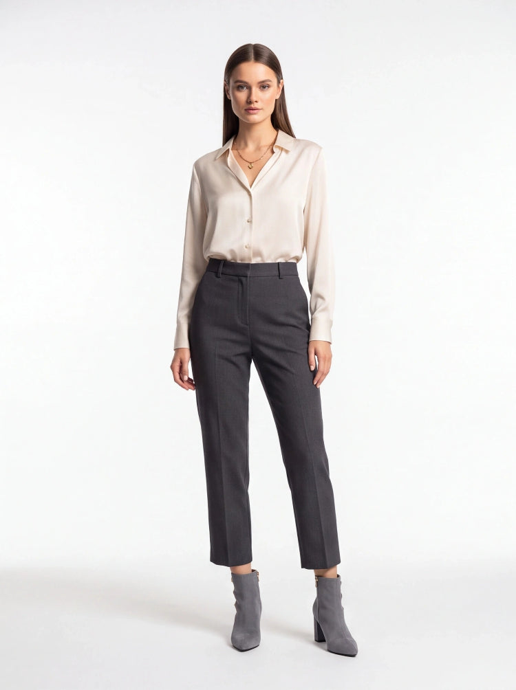 A woman modeling a light beige long-sleeve button-up shirt and dark charcoal gray cropped trousers, paired with gray ankle boots, against a plain white studio background.
