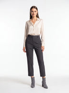 A woman modeling a light beige long-sleeve button-up shirt and dark charcoal gray cropped trousers, paired with gray ankle boots, against a plain white studio background.