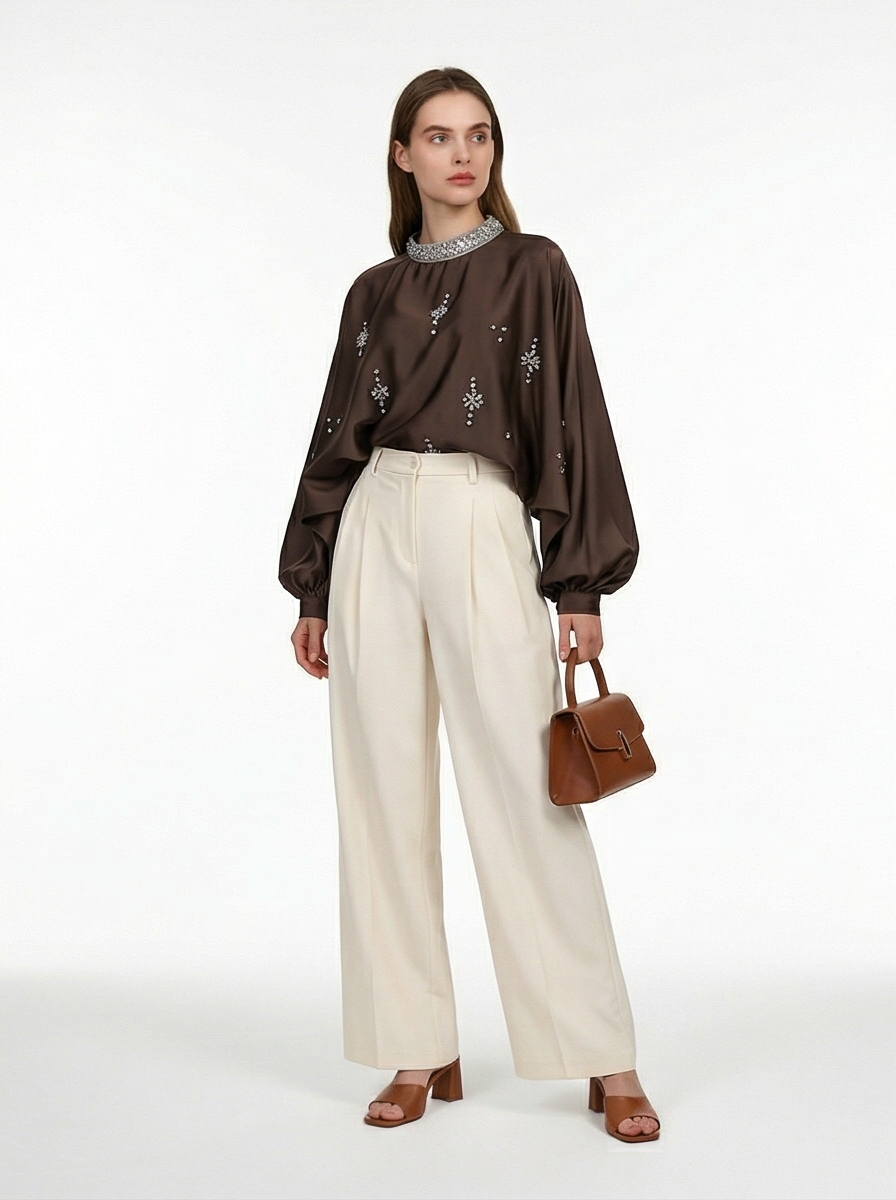 Woman wearing a brown blouse with floral embroidery and beige pants, holding a brown handbag on a white background.