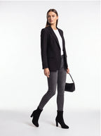 A woman modeling a black blazer, white top, dark gray skinny jeans, black ankle boots, and holding a black handbag against a plain white studio background.