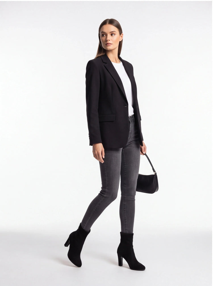 A woman modeling a black blazer, white top, dark gray skinny jeans, black ankle boots, and holding a black handbag against a plain white studio background.