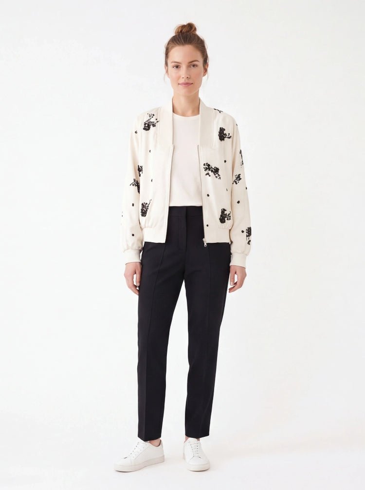 A model stands against a light gray studio background wearing a white sleeveless top with a small rectangular chest patch, a black knee-length skirt, a red and black patterned belt with gold hardware, and black pointed heels.