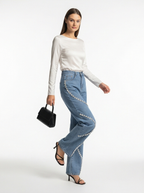 Woman wearing a white long-sleeve top and blue jeans with embellishments on a white background