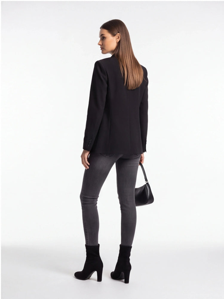A woman is shown from the back wearing a black blazer, dark gray skinny jeans, and black ankle boots, holding a small black handbag against a plain white studio background.
