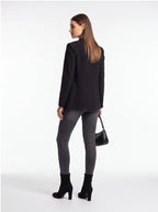 A woman is shown from the back wearing a black blazer, dark gray skinny jeans, and black ankle boots, holding a small black handbag against a plain white studio background.