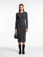 A woman stands against a plain light gray studio background wearing a fitted long-sleeve midi dress in dark charcoal gray, paired with black knee-high socks and a small black handbag.