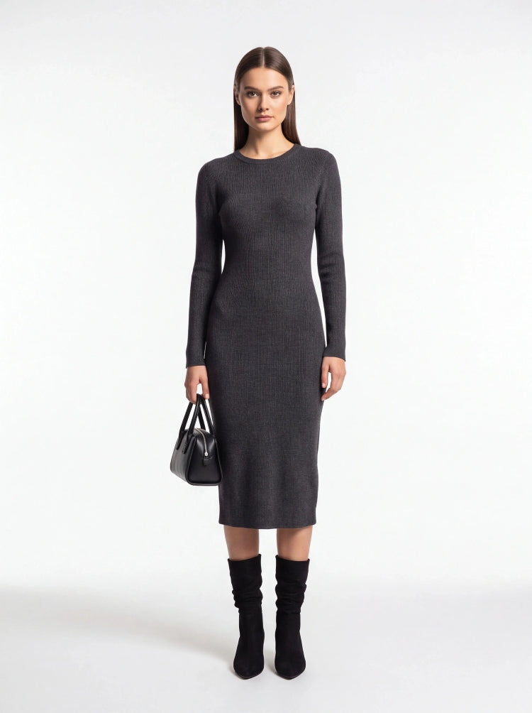 A woman stands against a plain light gray studio background wearing a fitted long-sleeve midi dress in dark charcoal gray, paired with black knee-high socks and a small black handbag.