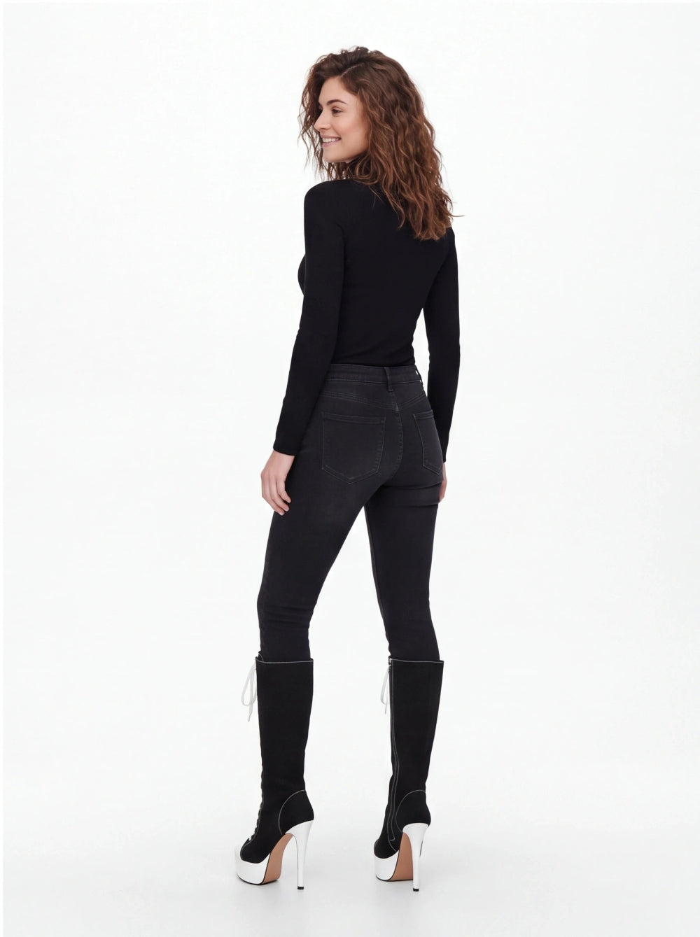 A woman shown from the back wearing a fitted black long-sleeve top, black skinny jeans, and black knee-high heeled boots with white platform soles, photographed against a clean white studio background.