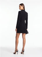A woman modeling a black long-sleeve mini blazer dress from the back, paired with black high-heeled sandals and holding a small black clutch, set against a plain white studio background.
