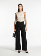 Woman wearing a beige sleeveless top and black wide-leg pants, holding a gray handbag on a white background.