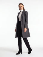 A woman modeling a long charcoal gray wool-blend coat over a white mock neck top, black skirt, black trousers, and black ankle boots, photographed against a seamless white studio background.