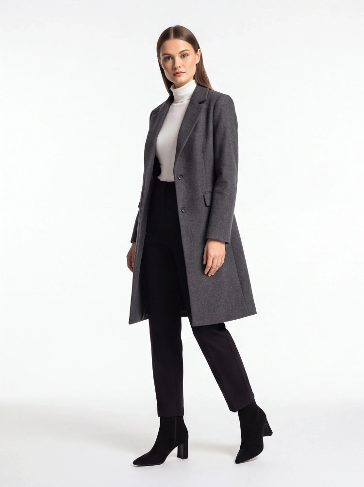 A woman modeling a long charcoal gray wool-blend coat over a white mock neck top, black skirt, black trousers, and black ankle boots, photographed against a seamless white studio background.
