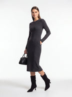 A woman modeling a dark gray long-sleeve knit midi dress with side pockets, paired with black ankle boots and a black handbag, against a plain white studio background.