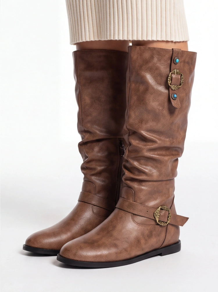 A pair of knee-high brown faux-leather boots with a slouchy fit, featuring decorative brass buckle straps and turquoise accents on the shaft, shown on a model wearing a cream ribbed skirt against a white studio background.